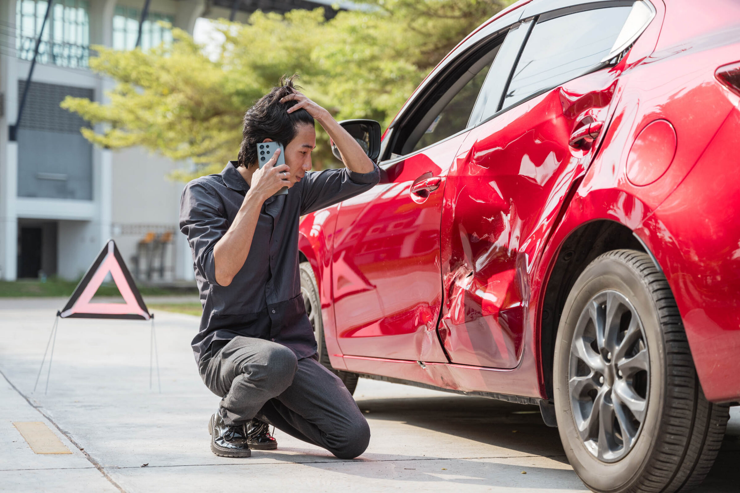 Worried man looks at his red car that has been in an accident. On the phone with car insurance, underinsured or uninsured driver.